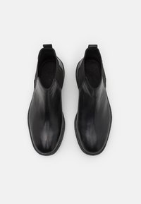 Botas de cuero negro hasta el tobillo con un diseño elegante, puntera redondeada, paneles laterales elásticos y tirador para facilitar el calzado. Suela texturizada visible.
