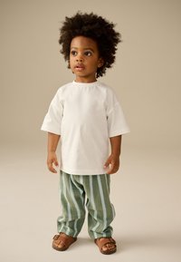 Camiseta blanca de manga corta, pantalones sueltos a rayas verdes y blancas, sandalias marrones con detalle de hebilla, tela texturizada, niño de pie frente a un fondo neutro.