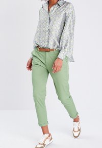 Chemise à manches longues à motifs verts, bleus et jaunes, manches retroussées. Portée avec un pantalon vert clair et des baskets décontractées.