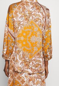 Blouse jaune et blanche à motifs cachemire et floraux, avec une coupe ample et des manches froncées, confectionnée en tissu doux.