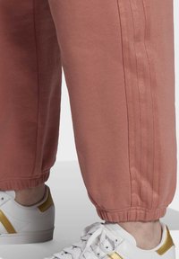 Rosa träningsoverallbyxor med elastiska muddar, med en mjuk textur och två vertikala ränder längs sidorna. I kombination med vita sneakers med guldaccenter.