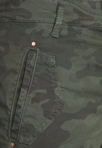 Tessuto verde a motivo mimetico con una grande tasca angolare, dotata di accessori color rame e dettagli di cucitura a contrasto.