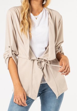 Donna che indossa una giacca beige aperta con maniche arrotolate e cintura annodata in vita sopra una maglietta bianca e jeans blu.