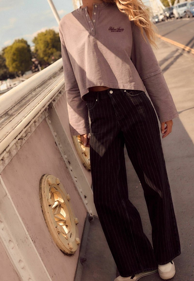 Femme portant un haut court lavande à manches longues et un pantalon large noir à fines rayures, debout près d'une rambarde par une journée ensoleillée.