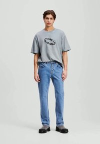 T-shirt de algodão cinza com uma impressão gráfica circular preta, combinado com jeans de denim azul claro. O modelo usa sapatos pretos, posando contra um fundo neutro.