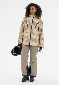 Beige camouflaged jacket met zwarte accenten, frontzakken en hoge kraag; gecombineerd met lichtkhaki broeken en zwarte skibotten.