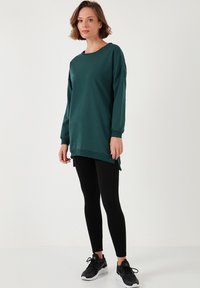 Zöld, hosszú ujjú pulóver kerek nyakkivágással, oldalvágásokkal, fekete leggings és fekete sportcipő fölött, semleges háttér előtt.