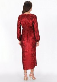 Frau, die ein langärmeliges, rotes Samt-Midikleid mit tailliertem Schnitt trägt und mit dem Rücken zum Betrachter vor einem schlichten weißen Hintergrund steht.