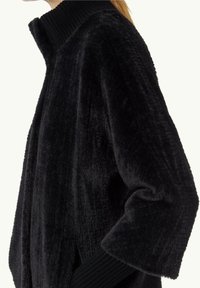 Vrouw in een zwarte, fluffy nepbontjas met een geribde hoge kraag en geribde manchetten, van opzij weergegeven.