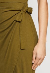 Détail d'une robe vert olive avec un lien à la taille, confectionnée dans un tissu doux au toucher lisse et au design drapé.