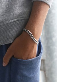 Zilveren armband met vergrendelde schakels, gedragen om een pols, Contrasterend met een grijze trui en blauwe broek. Gladde, glanzende oppervlak.
