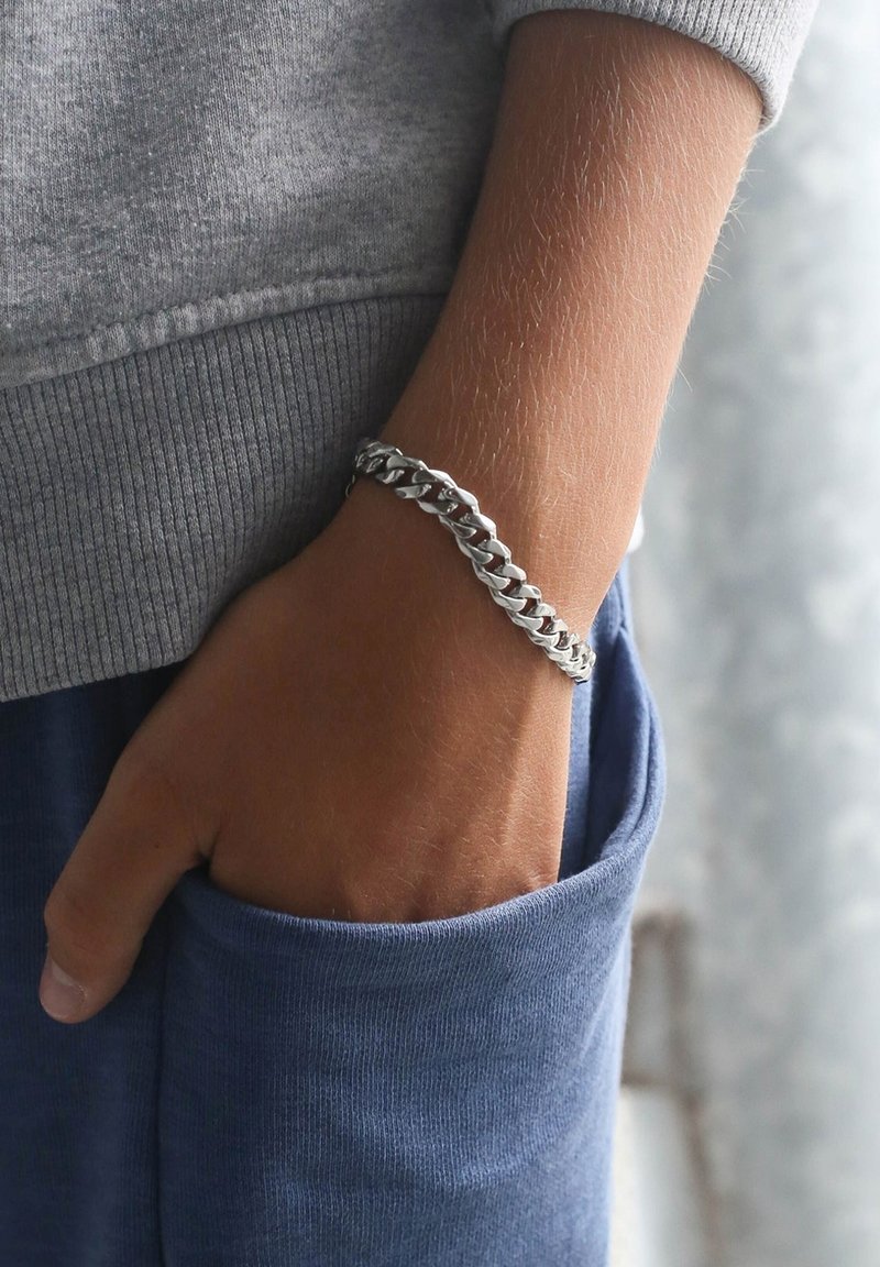 Zilveren armband met vergrendelde schakels, gedragen om een pols, Contrasterend met een grijze trui en blauwe broek. Gladde, glanzende oppervlak.
