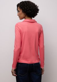 Polo corail à manches longues avec col, présentant une texture lisse et des accents rayés contrastants aux poignets, associée à un jean foncé.