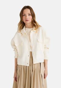 Cardigan blanc à boutons avec deux poches avant, poignets côtelés et coupe décontractée, associé à une jupe plissée beige clair.