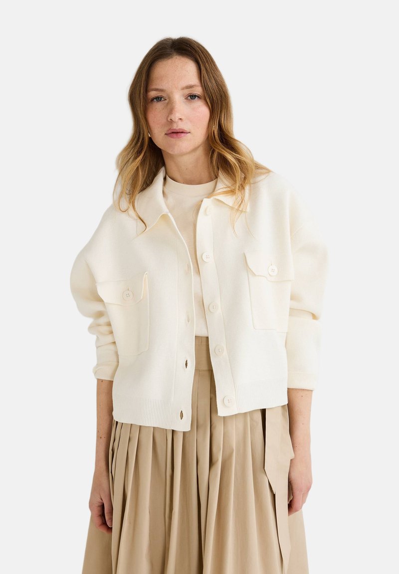 Cardigan blanc à boutons avec deux poches avant, poignets côtelés et coupe décontractée, associé à une jupe plissée beige clair.