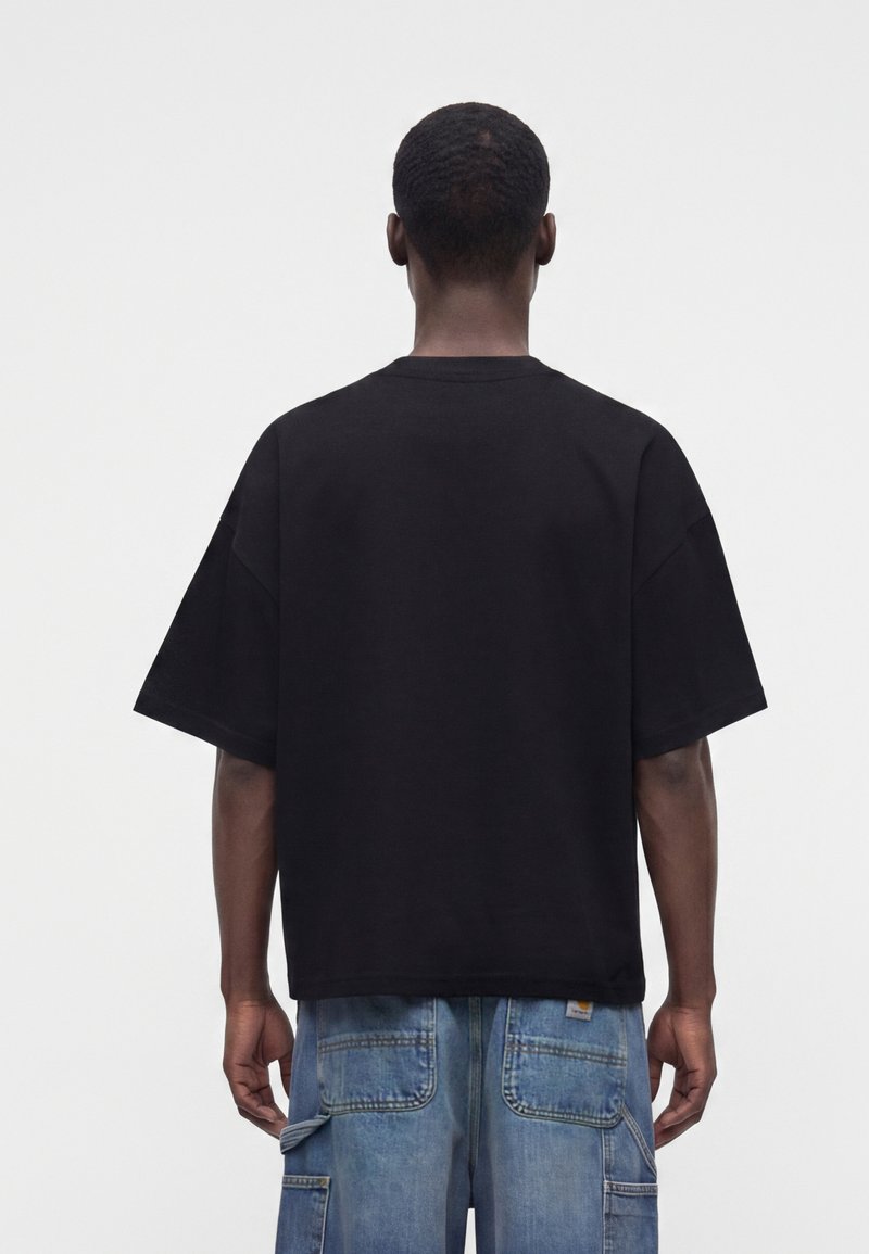 Mann von hinten gesehen, trägt ein lockeres schwarzes T-Shirt und blaue Denim-Cargohosen vor einem einfarbigen hellen Hintergrund.