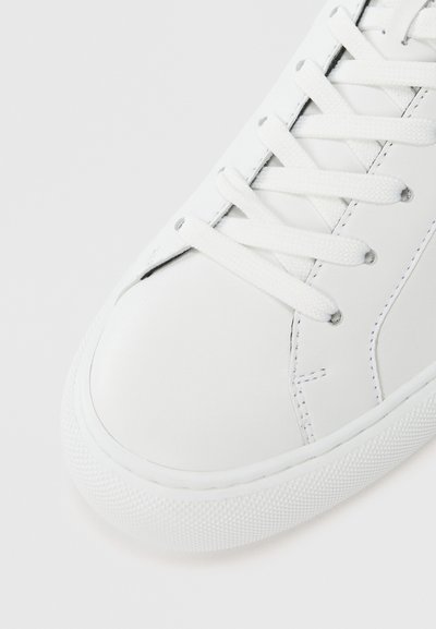 Close-up of a white leather sneaker's front with white laces crisscrossed through metal eyelets on a white textured sole.