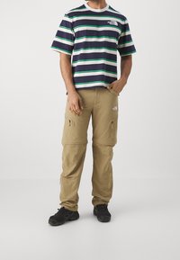 Mężczyzna ubrany w niebiesko-białą zieloną koszulkę w paski oraz beżowe spodnie cargo z zamkiem błyskawicznym, w czarnych butach turystycznych, stojący z jedną ręką w kieszeni.