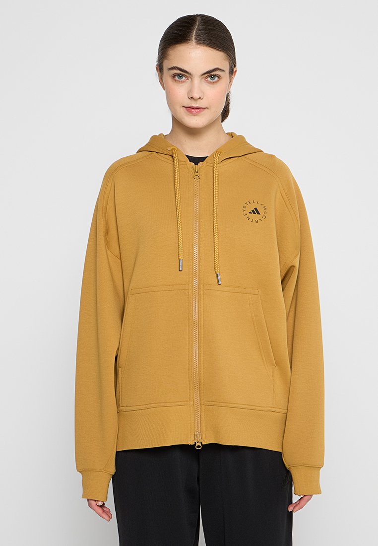 Sudadera amarilla con cremallera, fabricada en tejido suave, que presenta un logo en la parte frontal, dos bolsillos laterales y capucha ajustable con cordón.