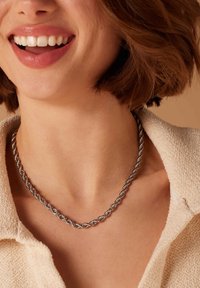 Zilveren gedraaide ketting met verweven schakel, zittend tegen de huid op de hals, gecombineerd met een textiel met een lichte kleur.