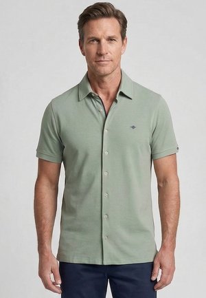 Man draagt een lichtgroen kortarmig polohemd met knopen en donkerblauwe broek, staand tegen een effen lichtgrijze achtergrond.