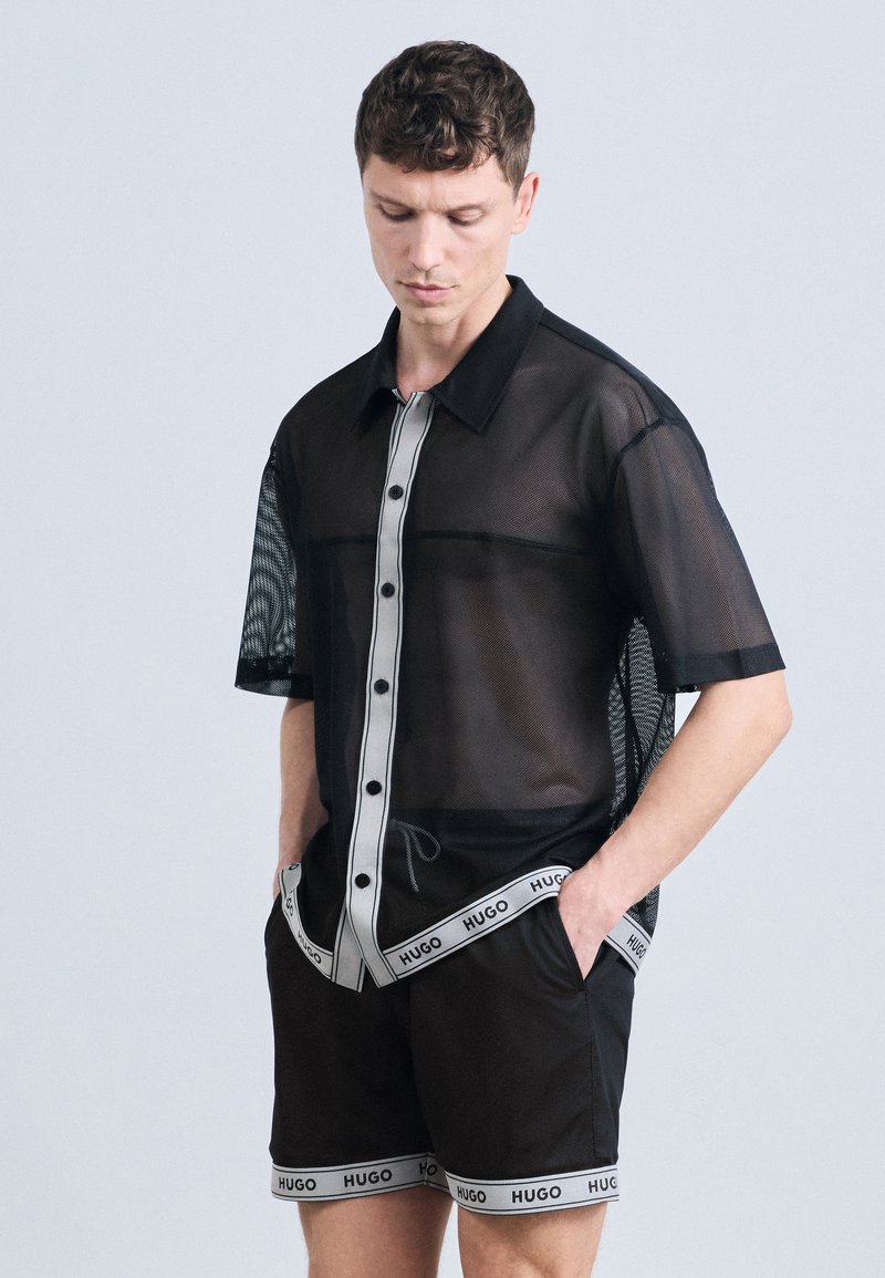 Άνδρας με μαύρο διαφανές πουκάμισο με κουμπιά και ταιριαστό σορτς με λευκή επένδυση, φέρει την ετικέτα "HUGO", στέκεται με τα χέρια στις τσέπες και κοιτάζει προς τα κάτω.