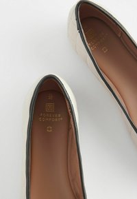 Weiße, gesteppte Ballerinas mit einem kontrastierenden schwarzen Rand und braunem Innenfutter, auf der Innensohle mit einem "Forever Comfort"-Logo.