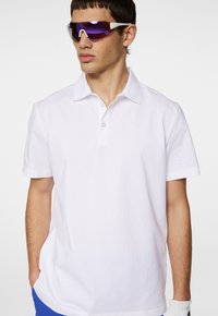 Vit pikétröja med klassisk krage och tre knappar; modellen bär lila tonade solglasögon och en vit handske.