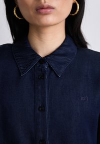 Camicia in denim blu scuro con colletto a punta, bottoni neri e un logo ricamato in modo discreto sul lato sinistro. Texture liscia.