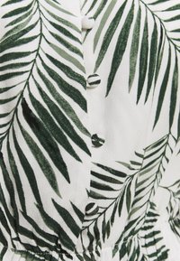 Witte stof met een donkergroene palmbladprint, knoopaccenten en een gathered textuur rond de taille.