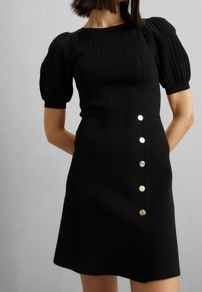 Femme portant un haut noir à côtes avec manches courtes bouffantes et une jupe noire ornée de cinq boutons métalliques verticaux sur le côté.