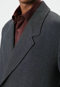 Grijze visgraatjas met een verzonken kraag en een gestructureerde afwerking, gedragen over een bordeauxrode overhemd met knoopsluiting en een standaard kraag.