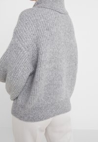 Harmaa neulottu villapaita korkealla kauluksella, jossa on resorimainen tekstuuri ja väljä istuvuus. Näkyvät saumat ja rennot hihat lisäävät mukavuutta.