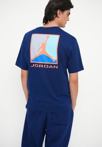 Jūras zilas kokvilnas T-kreklis ar lielu oranžu un zilu Jordan logotipa grafiku un tekstu aizmugurē, ar apaļu kakla izgriezumu un īsām piedurknēm.