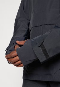 Chaqueta gris oscuro resistente al agua con mangas largas, que presenta cierres de Velcro y una textura suave. Vista en primer plano de manos sosteniendo la manga.