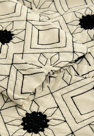 Prošívaná látka s krémovým pozadím a černými geometrickými vzory a květinovými motivy. Texturová černá nit dodává hloubku.