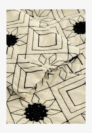 Gesteppten Stoff mit einem cremefarbenen Hintergrund, der mit schwarzen geometrischen Mustern und floralen Motiven versehen ist. Strukturierte schwarze Nähte verleihen Tiefe.