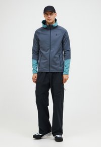 Grå softshell-jakke med teal ærmer, frontlynlås og sidelommer, parret med sorte cargo-bukser og en sort kasket.