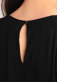 Indumento nero con un'apertura a chiave sulla schiena, fissata da un bottone nero. Il materiale sembra morbido e leggermente testurizzato.