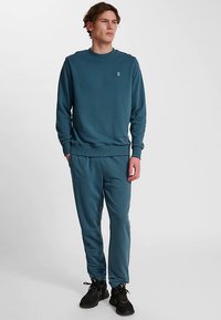 Türkisfarbener Pullover und passende Jogginghosen aus weichem Stoff. Verfügt über gerippte Bündchen und Bund sowie ein kleines Logo auf der Brust. Schwarze Turnschuhe getragen.