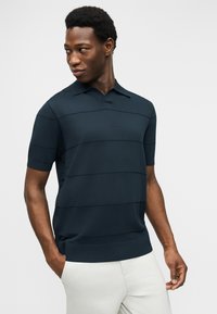 Άνδρας που φοράει σκουρό μπλε κοντομάνικη πόλο μπλούζα με οριζόντιες ρίγες και ανοιχτόχρωμο παντελόνι, με τα χέρια στις τσέπες, κοιτάζει προς τα αριστερά του.