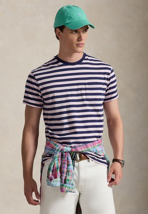 Μπλουζάκι με ριγέ κοντά μανίκια σε ναυτικό μπλε και ανοιχτό ροζ, με τσέπη στο στήθος. Λευκό τζιν και πουκάμισο καρό δεμένο στη μέση. Τζελ πράσινο καπέλο.