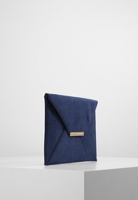 Clutch in finta pelle scamosciata blu navy con design geometrico, accento in metallo dorato, patta triangolare e dettagli cuciti sulla superficie.