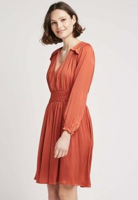 Robe couleur rouille avec un décolleté en V profond, des manches longues avec des poignets froncés et une taille smockée. Tissu doux et fluide avec une jupe évasée.