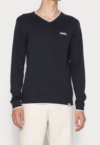 Mężczyzna ubrany w czarny sweter z długim rękawem, dekoltem w serek, ozdobiony białą lamówką i małym logo na piersi, w połączeniu z jasnobeżowymi spodniami.