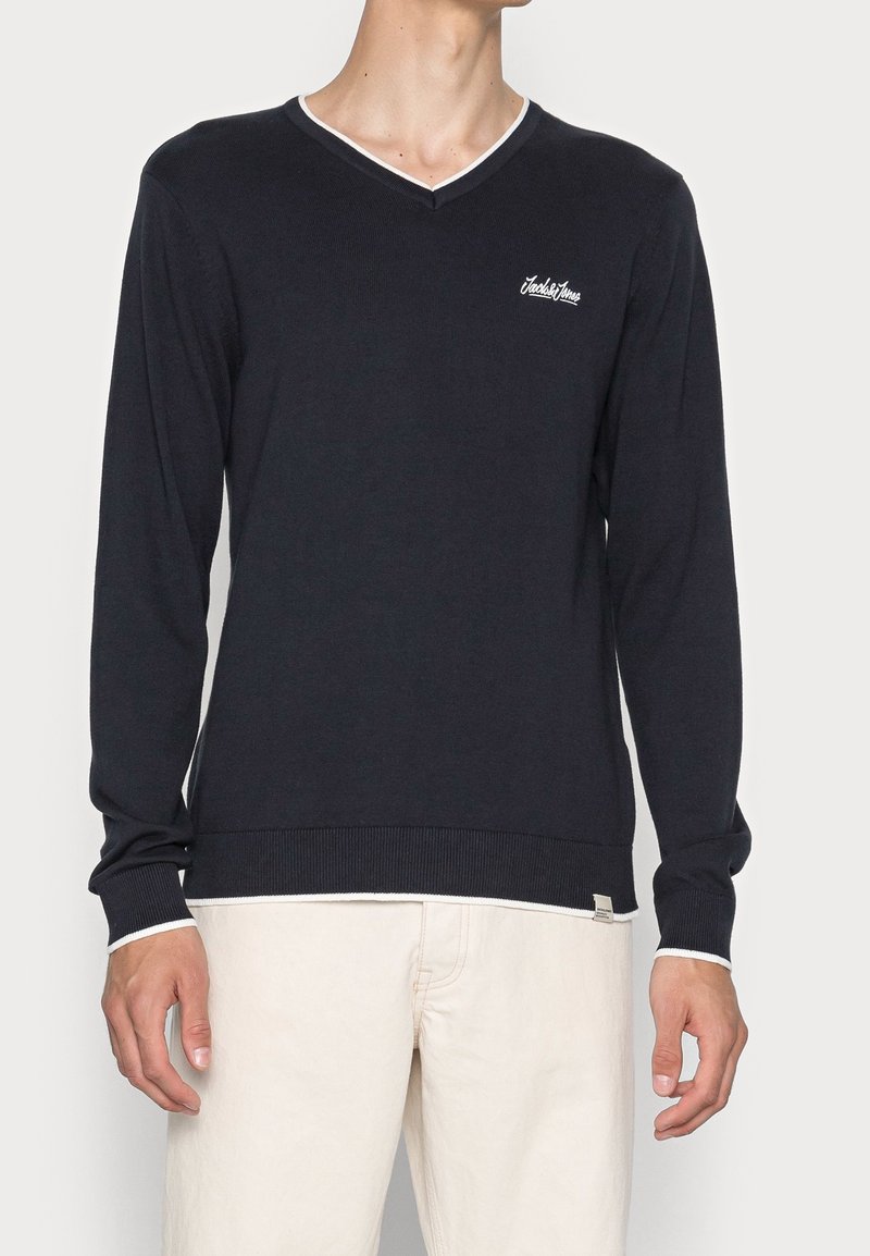 Mężczyzna ubrany w czarny sweter z długim rękawem, dekoltem w serek, ozdobiony białą lamówką i małym logo na piersi, w połączeniu z jasnobeżowymi spodniami.