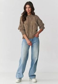 Brązowy sweter z teksturowanym splotem, z prążkowanymi mankietami, w połączeniu z jasnoniebieskimi, luźnymi dżinsami. Model stoi w białych trampkach na jednolitym tle.