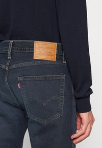 Jeans de denim azul escuro com um remendo de couro texturizado na parte de trás, apresentando um logótipo cosido. O bolso de trás tem um design curvado.