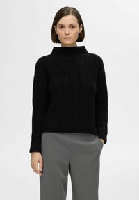 Pull en maille côtelée noir avec un col haut et des manches larges. Porté avec un pantalon gris à coupe ample. Tissu lisse et texturé.