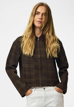 Femme aux longs cheveux blonds portant une veste zippée à carreaux marron foncé et un pantalon blanc, mains dans les poches, expression neutre, debout devant un fond uni.